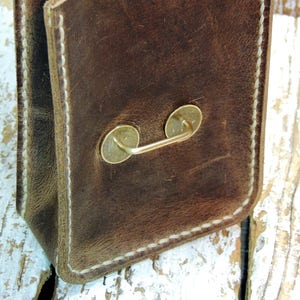May include: A brown leather bag with a white stitched border and a brass handle. The bag is sitting on a white wooden surface.