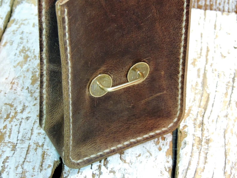 May include: A brown leather bag with a white stitched border and a brass handle. The bag is sitting on a white wooden surface.