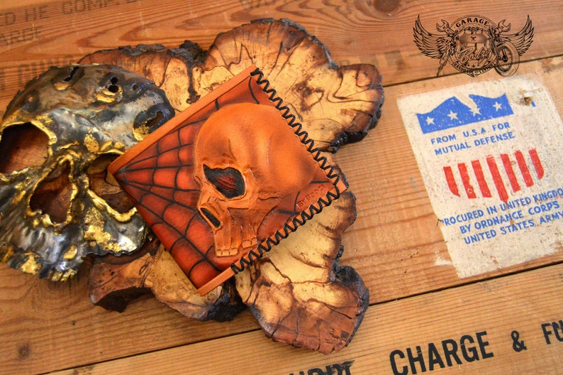 May include: A brown leather wallet with a skull and spiderweb design. The wallet is stitched with black thread and has a distressed look.