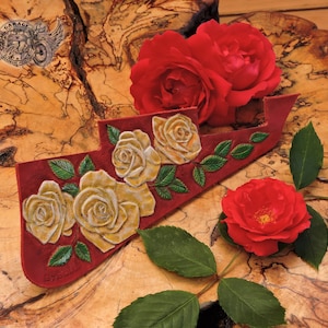 Puede incluir: Una púa de guitarra de cuero rojo con un diseño tallado de rosas blancas y hojas verdes. La púa está sobre una superficie de madera con rosas rojas.