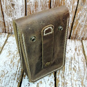 May include: A brown leather pouch with a white stitch detail and a flap closure. The pouch has a strap with a metal button and two metal rivets.