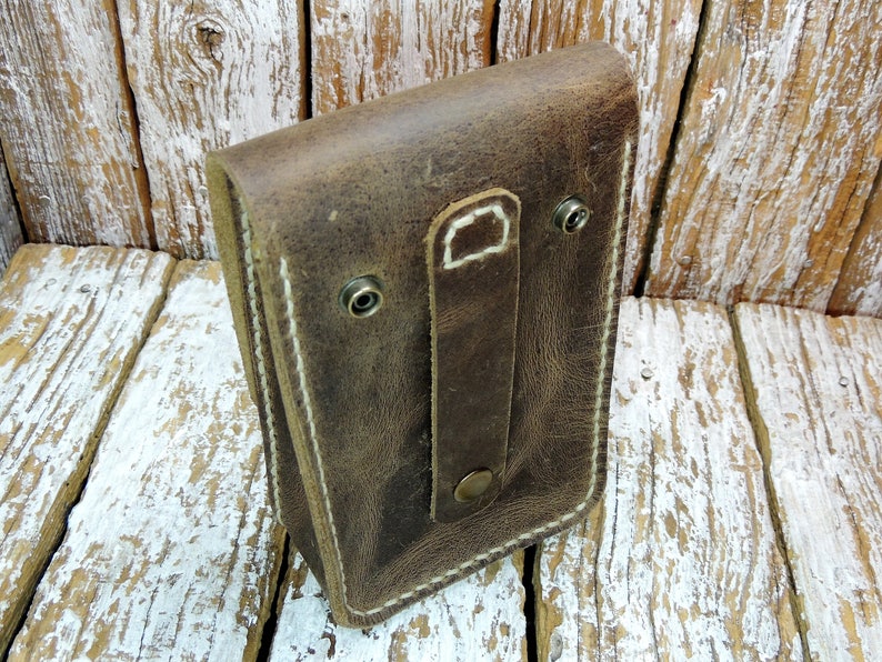 May include: A brown leather pouch with a white stitch detail and a flap closure. The pouch has a strap with a metal button and two metal rivets.