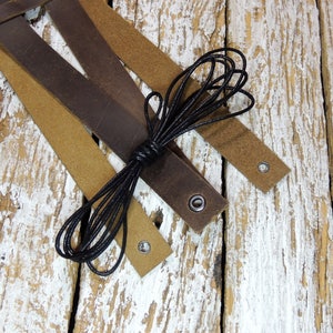 May include: Three leather straps in brown and tan with black cord and metal eyelets. The straps are arranged on a whitewashed wooden surface.
