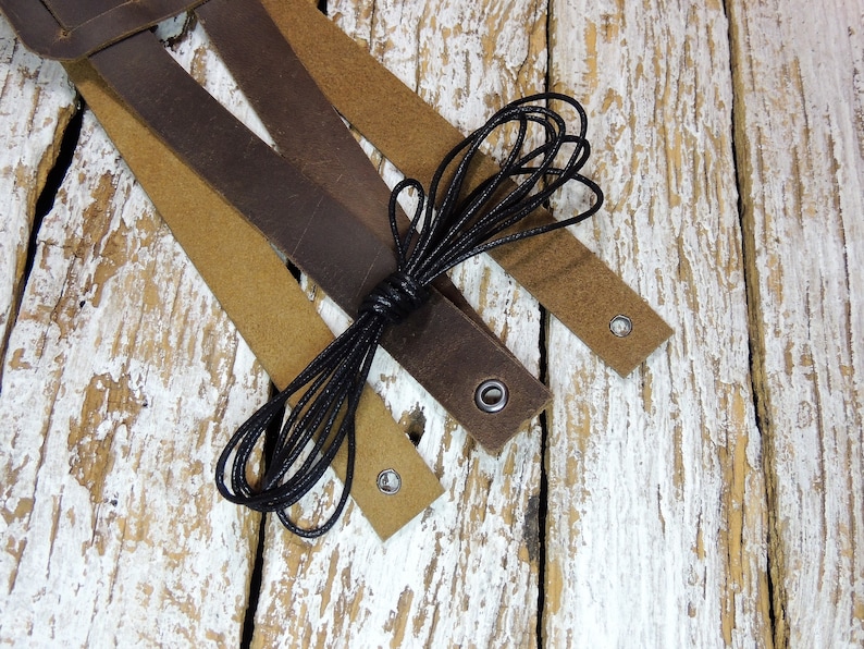 May include: Three leather straps in brown and tan with black cord and metal eyelets. The straps are arranged on a whitewashed wooden surface.