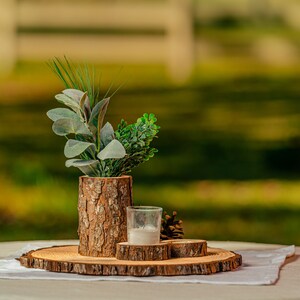 May include: A rustic centerpiece arrangement on a round wooden slab. It features a bark-covered vase with greenery, a small glass candle holder, and a pine cone. The arrangement sits on a white cloth, with a blurred outdoor background.