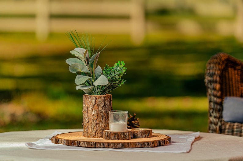May include: A rustic centerpiece arrangement on a round wooden slab. It features a bark-covered vase with greenery, a small glass candle holder, and a pine cone. The arrangement sits on a white cloth, with a blurred outdoor background.