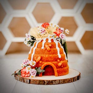 Puede incluir: Un pastel con forma de colmena decorado con glaseado blanco y un arreglo floral de flores rosas, amarillas y blancas. El pastel est&aacute; sobre una rodaja de madera.