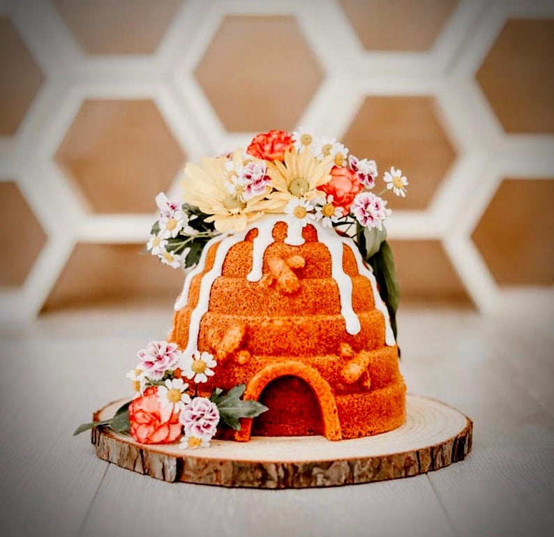 Puede incluir: Un pastel con forma de colmena decorado con glaseado blanco y un arreglo floral de flores rosas, amarillas y blancas. El pastel est&aacute; sobre una rodaja de madera.