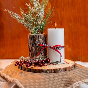 May include: A white pillar candle with a red and black checkered ribbon and a burning flame. A rustic wooden vase holds greenery and white flowers. The arrangement sits on a wooden slice with red berries.