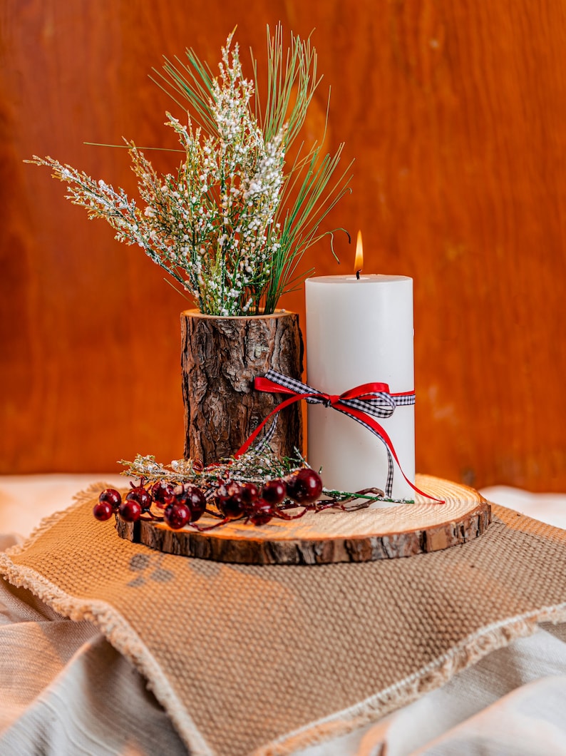 May include: A white pillar candle with a red and black checkered ribbon and a burning flame. A rustic wooden vase holds greenery and white flowers. The arrangement sits on a wooden slice with red berries.