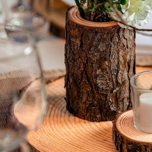 May include: A wooden log slice with a smaller log slice on top, used as a decorative centerpiece. A small glass candle holder sits on the top slice, and a potted plant with white flowers sits on the bottom slice.