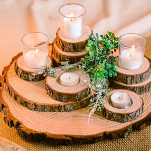 May include: A rustic wooden candle holder arrangement. The centerpiece features multiple levels of wood slices, each holding a lit candle in a glass votive or a small tea light. Greenery adds a natural touch to the display.