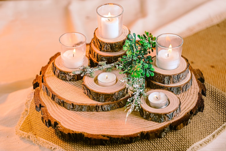 May include: A rustic wooden candle holder arrangement. The centerpiece features multiple levels of wood slices, each holding a lit candle in a glass votive or a small tea light. Greenery adds a natural touch to the display.