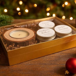 May include: A gold gift box contains a candle in a wooden holder and three small scented candles. The candles are surrounded by decorative filler. Red Christmas ornaments and pinecones are scattered around the box, with a blurred background of Christmas lights.