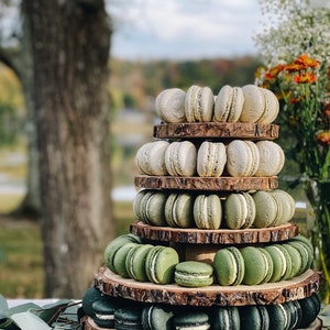 May include: A tiered display of macarons in shades of green and white, arranged on rustic wooden slices. The macarons are stacked to create a visually appealing dessert presentation. The background features a blurred outdoor setting with trees and flowers.