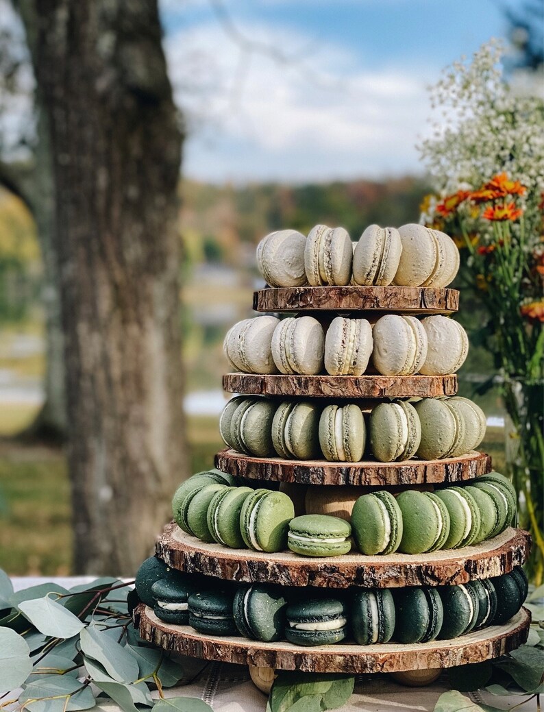 May include: A tiered display of macarons in shades of green and white, arranged on rustic wooden slices. The macarons are stacked to create a visually appealing dessert presentation. The background features a blurred outdoor setting with trees and flowers.