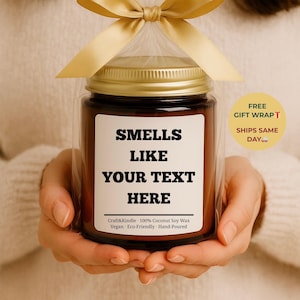 May include: A brown glass jar candle with a gold lid, wrapped in clear cellophane and tied with a gold ribbon. The label reads "SMELLS LIKE YOUR TEXT HERE". The candle is made with coconut soy wax and is vegan and eco-friendly.