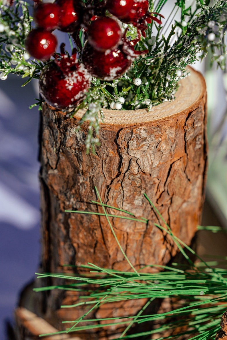 May include: A rustic wooden vase with a textured bark exterior. The vase holds a festive arrangement of red berries, green sprigs, and white accents, creating a natural and seasonal display. The vase is cylindrical in shape.