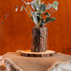 May include: A rustic wooden vase made from a tree trunk, holding green and gray artificial plants and branches. The vase sits on a wooden slice, displayed on a tan fabric with a wooden background.