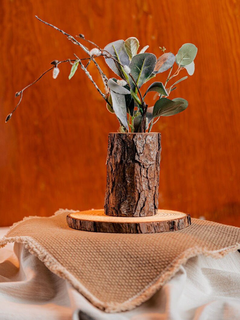 May include: A rustic wooden vase made from a tree trunk, holding green and gray artificial plants and branches. The vase sits on a wooden slice, displayed on a tan fabric with a wooden background.