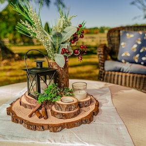 May include: A rustic centerpiece on a round table. It features a black lantern, candles, and decorative wood slices. A vase with greenery and berries adds color. The table is set with a white runner, and a wicker chair is in the background.