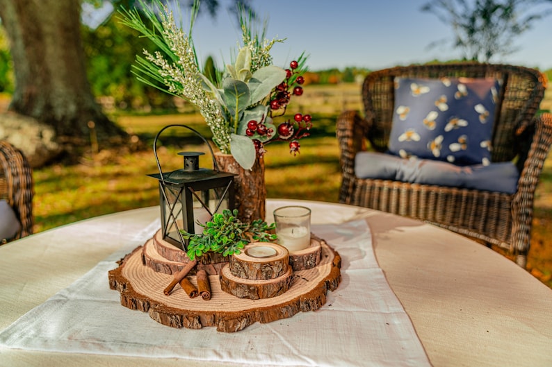 May include: A rustic centerpiece on a round table. It features a black lantern, candles, and decorative wood slices. A vase with greenery and berries adds color. The table is set with a white runner, and a wicker chair is in the background.