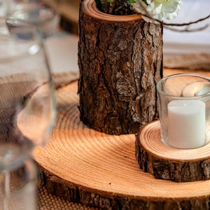 Puede incluir: Una rodaja de tronco de madera con una rodaja de tronco de madera m&aacute;s peque&ntilde;a en la parte superior. Un portavelas de vidrio con una vela blanca se coloca sobre la rodaja de tronco de madera m&aacute;s peque&ntilde;a. Una peque&ntilde;a planta en maceta con hojas verdes y flores blancas se coloca sobre la rodaja de tronco de madera m&aacute;s grande.