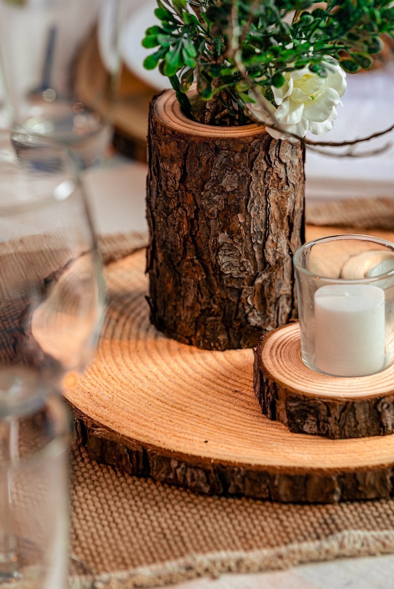 Puede incluir: Una rodaja de tronco de madera con una rodaja de tronco de madera m&aacute;s peque&ntilde;a en la parte superior. Un portavelas de vidrio con una vela blanca se coloca sobre la rodaja de tronco de madera m&aacute;s peque&ntilde;a. Una peque&ntilde;a planta en maceta con hojas verdes y flores blancas se coloca sobre la rodaja de tronco de madera m&aacute;s grande.
