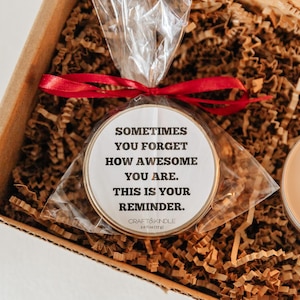 May include: A gift box with a candle and a round, packaged item. The round item has text that reads, "Sometimes you forget how awesome you are. This is your reminder." The box is filled with brown paper and tied with a red ribbon.