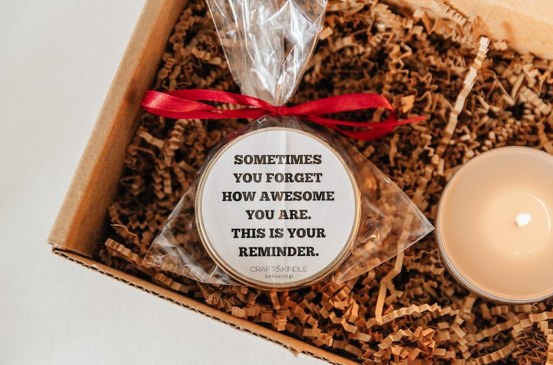 May include: A gift box with a candle and a round, packaged item. The round item has text that reads, "Sometimes you forget how awesome you are. This is your reminder." The box is filled with brown paper and tied with a red ribbon.