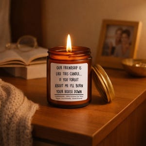 May include: A lit brown glass candle jar with a gold lid, sitting on a wooden surface. The candle has a white label with black text that reads, "OUR FRIENDSHIP IS LIKE THIS CANDLE... IF YOU FORGET ABOUT ME I'LL BURN YOUR HOUSE DOWN."