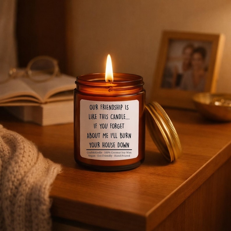 May include: A lit brown glass candle jar with a gold lid, sitting on a wooden surface. The candle has a white label with black text that reads, "OUR FRIENDSHIP IS LIKE THIS CANDLE... IF YOU FORGET ABOUT ME I'LL BURN YOUR HOUSE DOWN."