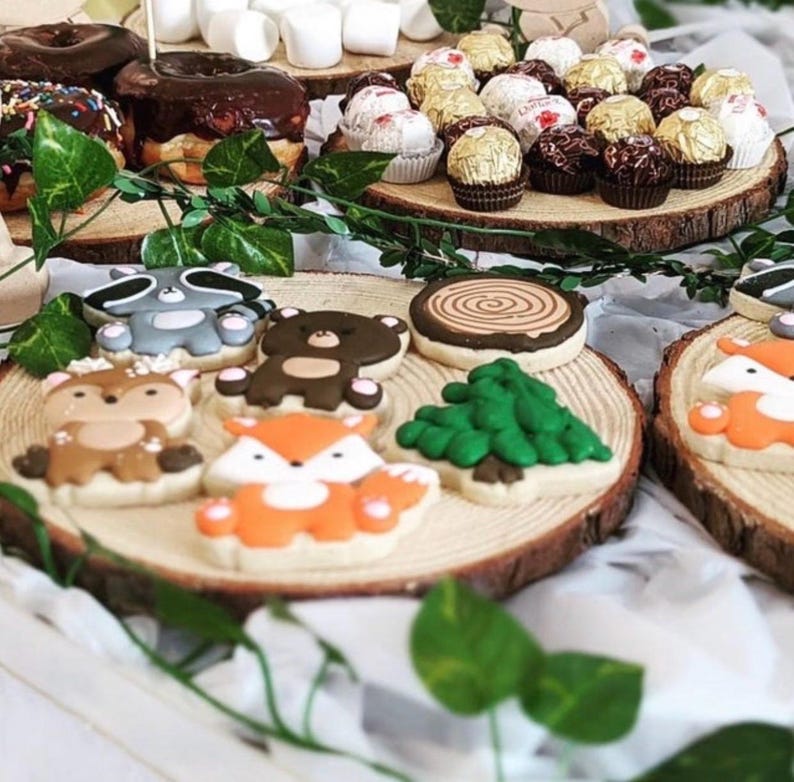 Puede incluir: Un plato de madera con una variedad de galletas de az&uacute;car decoradas con animales del bosque, incluyendo un mapache, un oso, un ciervo, un zorro y un pino. Las galletas est&aacute;n dispuestas en un plato de madera con un dise&ntilde;o de tronco de &aacute;rbol.