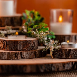 May include: A rustic candle display featuring wooden slices of varying sizes. Tealight candles and pillar candles in glass holders are arranged on the wooden platforms. Greenery and small white berries add a natural touch to the warm, inviting scene.