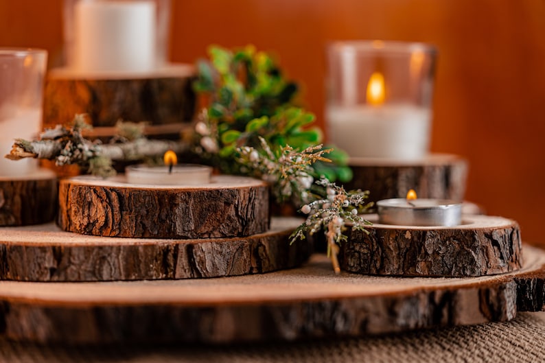 May include: A rustic candle display featuring wooden slices of varying sizes. Tealight candles and pillar candles in glass holders are arranged on the wooden platforms. Greenery and small white berries add a natural touch to the warm, inviting scene.