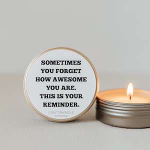 May include: A lit candle in a small, round, silver tin next to a matching tin with a white label. The label reads "Sometimes you forget how awesome you are. This is your reminder."