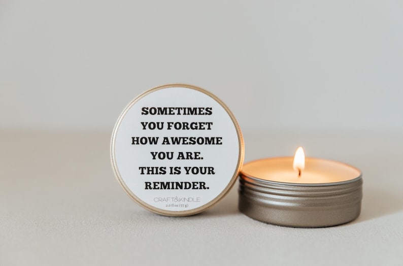 May include: A lit candle in a small, round, silver tin next to a matching tin with a white label. The label reads "Sometimes you forget how awesome you are. This is your reminder."