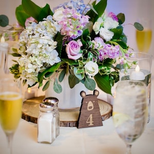 May include: A wooden bear table number with the number 4 on it. The table number is sitting on a wooden slice. A bouquet of purple, white, and blue flowers is in the background.