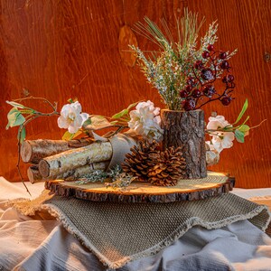 May include: A rustic centerpiece featuring a wooden log vase filled with greenery and red berries. White flowers and birch logs are arranged on a wooden slice, with a pine cone and burlap accents. The arrangement sits on a textured fabric.