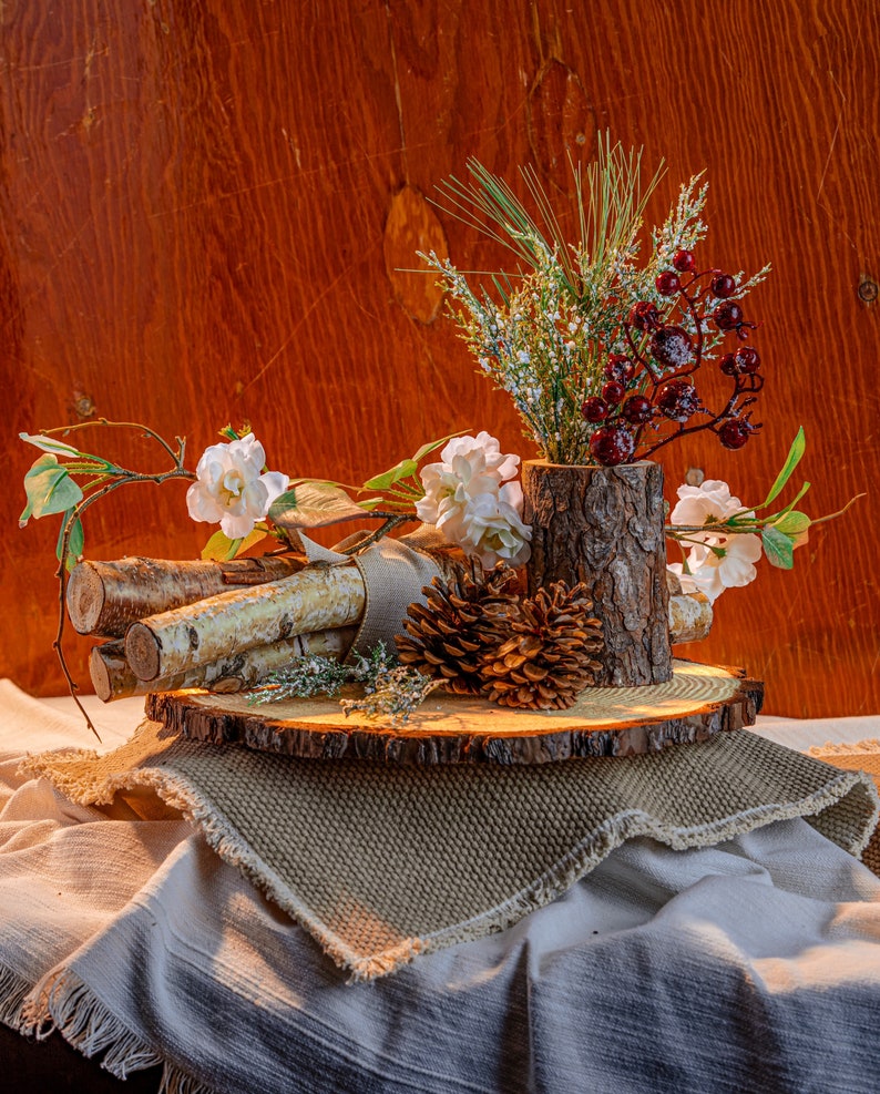 May include: A rustic centerpiece featuring a wooden log vase filled with greenery and red berries. White flowers and birch logs are arranged on a wooden slice, with a pine cone and burlap accents. The arrangement sits on a textured fabric.