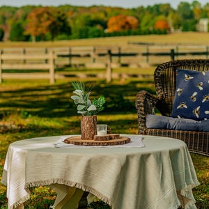 May include: An outdoor scene featuring a wicker rocking chair and a small round table. The chair has a blue cushion and a decorative pillow with a bee pattern. The table is covered with a fringed tablecloth and a wooden tray with a candle and greenery.