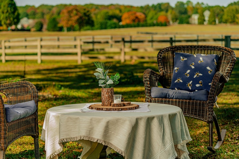 May include: An outdoor scene featuring a wicker rocking chair and a small round table. The chair has a blue cushion and a decorative pillow with a bee pattern. The table is covered with a fringed tablecloth and a wooden tray with a candle and greenery.