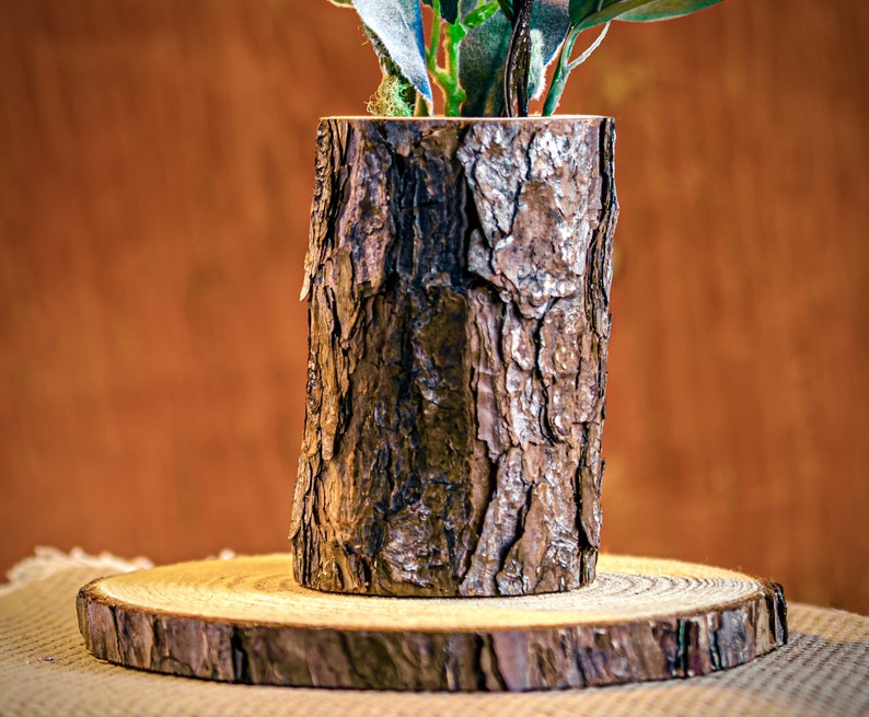 May include: A cylindrical wooden vase with a rough, textured bark exterior, holding green foliage. The vase sits on a round wooden base, showcasing a natural, rustic aesthetic. The vase is approximately 6 inches tall.