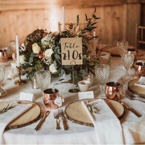 May include: A rustic wedding table setting with a centerpiece of greenery and flowers. The table is set with copper mugs, wooden chargers, white napkins, and a handwritten table number card that says "Table 110".