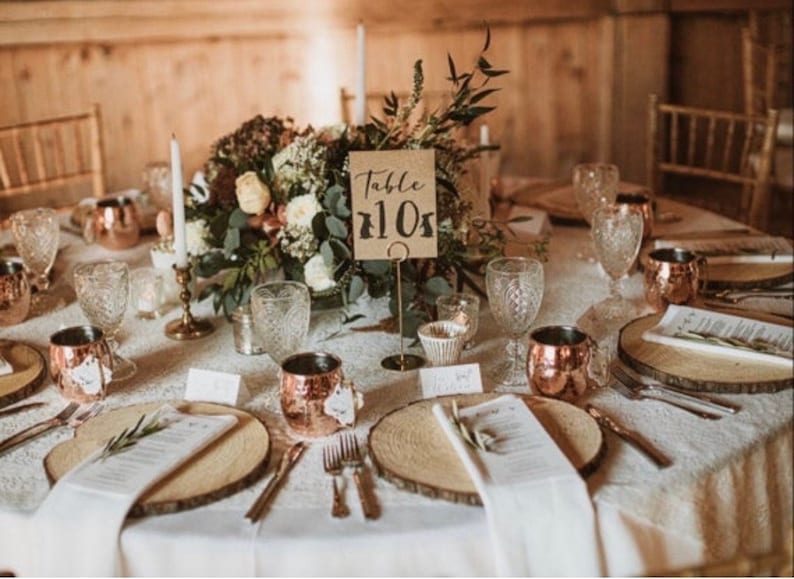 May include: A rustic wedding table setting with a centerpiece of greenery and flowers. The table is set with copper mugs, wooden chargers, white napkins, and a handwritten table number card that says "Table 110".