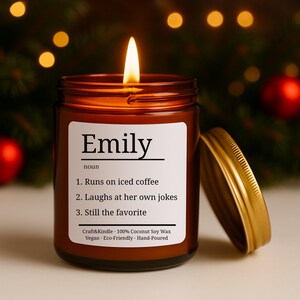 Puede incluir: Una vela encendida en un frasco de vidrio marr&oacute;n con tapa dorada. La etiqueta dice "Emily" seguida de una definici&oacute;n: "Corre con caf&eacute; helado, Se r&iacute;e de sus propios chistes, Sigue siendo la favorita". La vela est&aacute; vertida a mano y hecha con cera de soja de coco.
