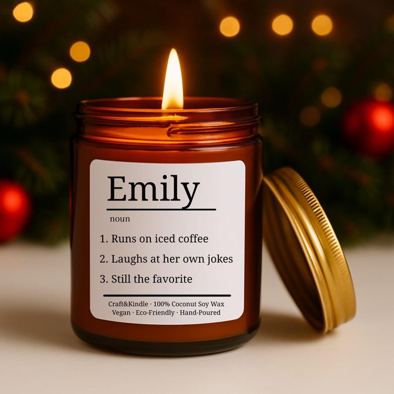 Puede incluir: Una vela encendida en un frasco de vidrio marr&oacute;n con tapa dorada. La etiqueta dice "Emily" seguida de una definici&oacute;n: "Corre con caf&eacute; helado, Se r&iacute;e de sus propios chistes, Sigue siendo la favorita". La vela est&aacute; vertida a mano y hecha con cera de soja de coco.