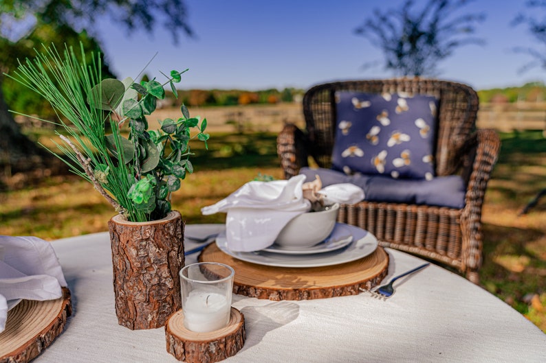 May include: Outdoor dining table setting with a white tablecloth, wooden chargers, and a centerpiece of greenery in a wood vase. Includes white plates, napkins, and a candle. A wicker chair with a bee-patterned pillow is in the background.