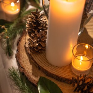 May include: A warm, inviting scene featuring a large white pillar candle and a small votive candle, both lit. Pine cones and greenery are arranged on a wooden slice, creating a cozy, natural aesthetic. A glass vase filled with pine cones is also visible.