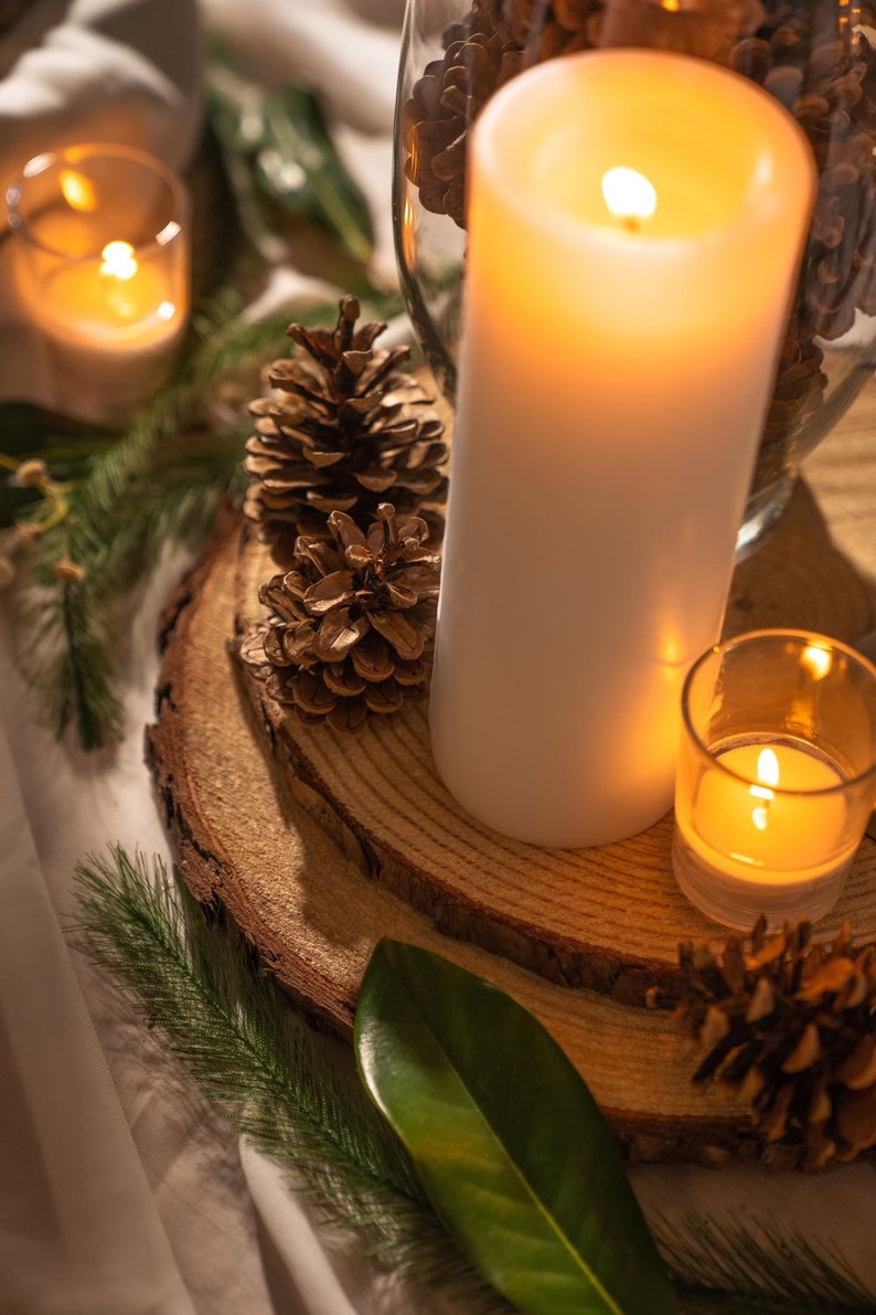 May include: A warm, inviting scene featuring a large white pillar candle and a small votive candle, both lit. Pine cones and greenery are arranged on a wooden slice, creating a cozy, natural aesthetic. A glass vase filled with pine cones is also visible.